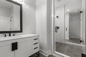 A modern bathroom with a large framed mirror above a white countertop and sink, black fixtures, a glass-enclosed shower with white tile walls, and a gray tile floor.