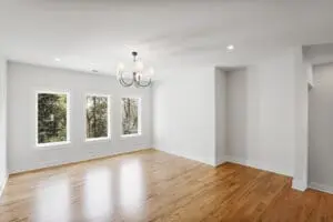 An empty room with hardwood floors, white walls, three windows, and a chandelier hanging from the ceiling.