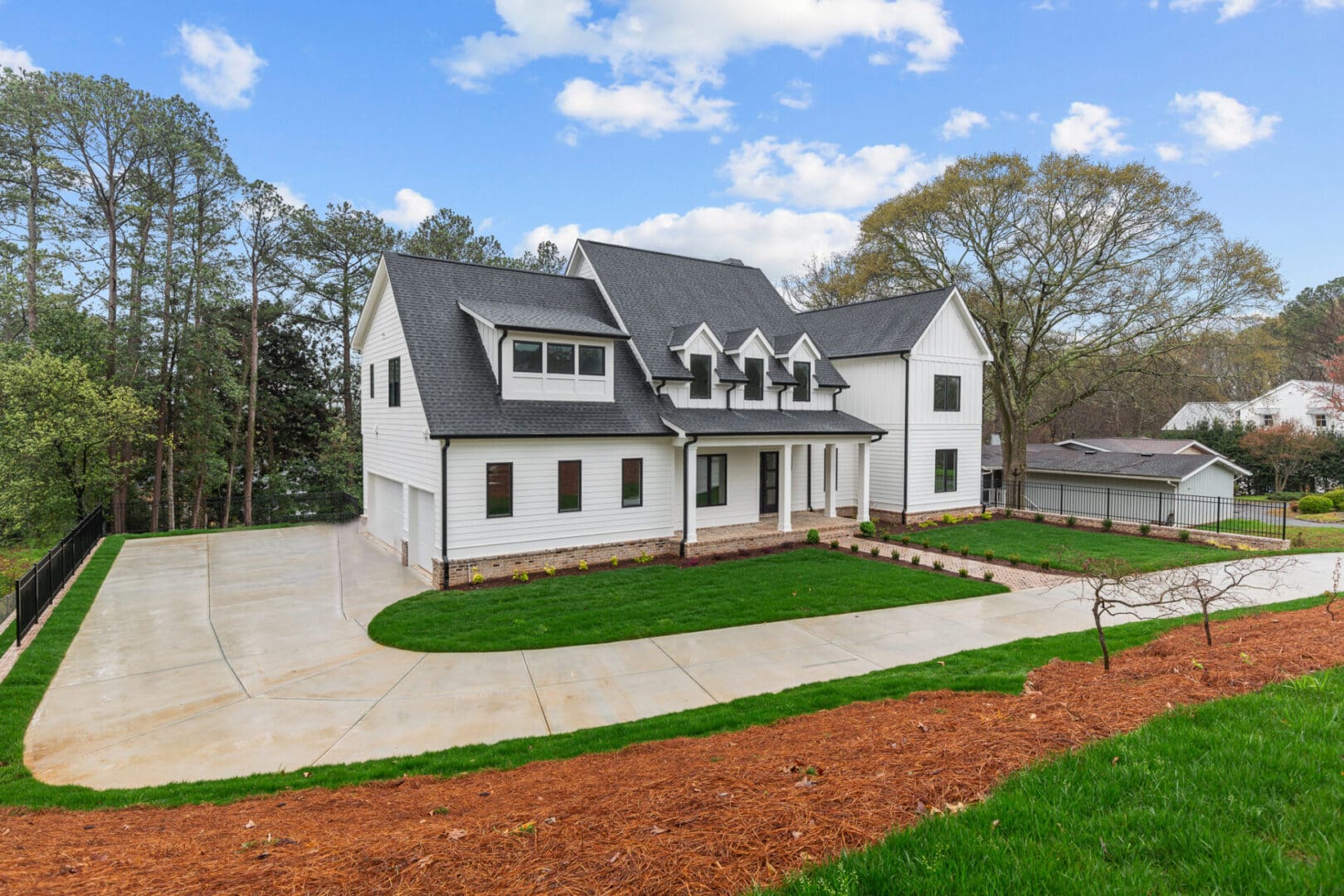 A modern two-story white house with black roofing, a three-car garage, and a spacious driveway surrounded by well-maintained lawns and trees under a partly cloudy sky.