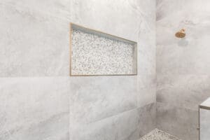 A minimalist bathroom shower with large light gray tiles, featuring a recessed shelf with mosaic tiles and a single bronze showerhead.