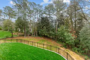 A backyard with a well-maintained green lawn, enclosed by a black metal fence, adjacent to a forested area with tall trees under a partly cloudy sky.