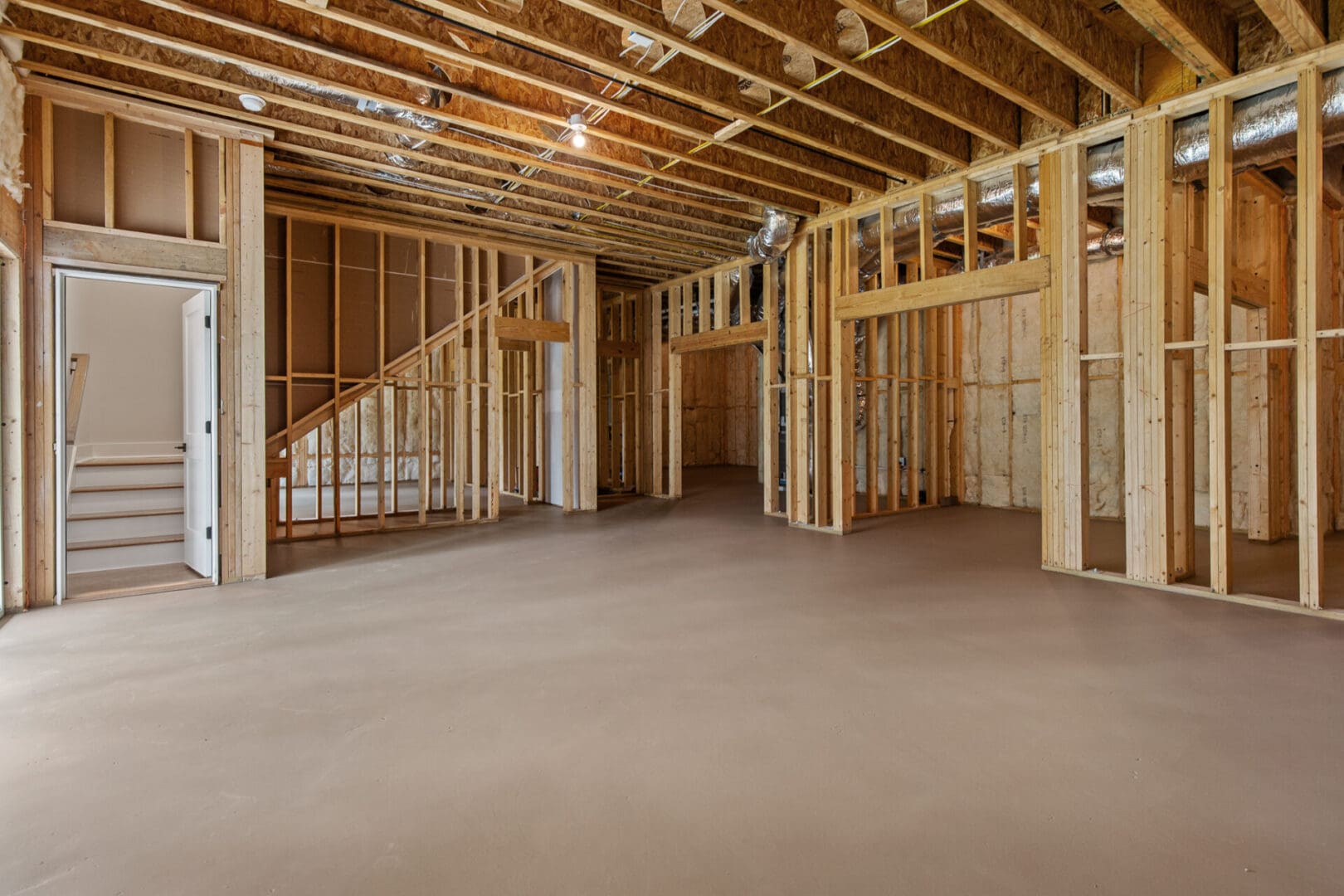 An unfinished basement with exposed wooden framing, unpainted drywall, and a concrete floor. A staircase leads upstairs, and ventilation ducts are visible on the ceiling. Natural light enters from the left.
