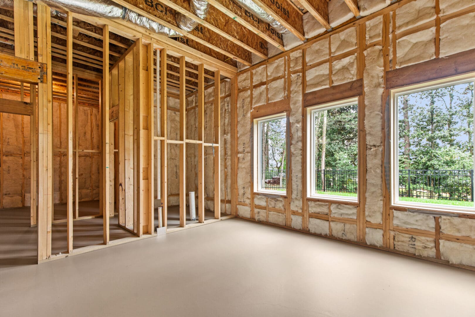 A partially constructed interior of a house showing wooden framing, wall insulation, and three large windows overlooking a green outdoor area.