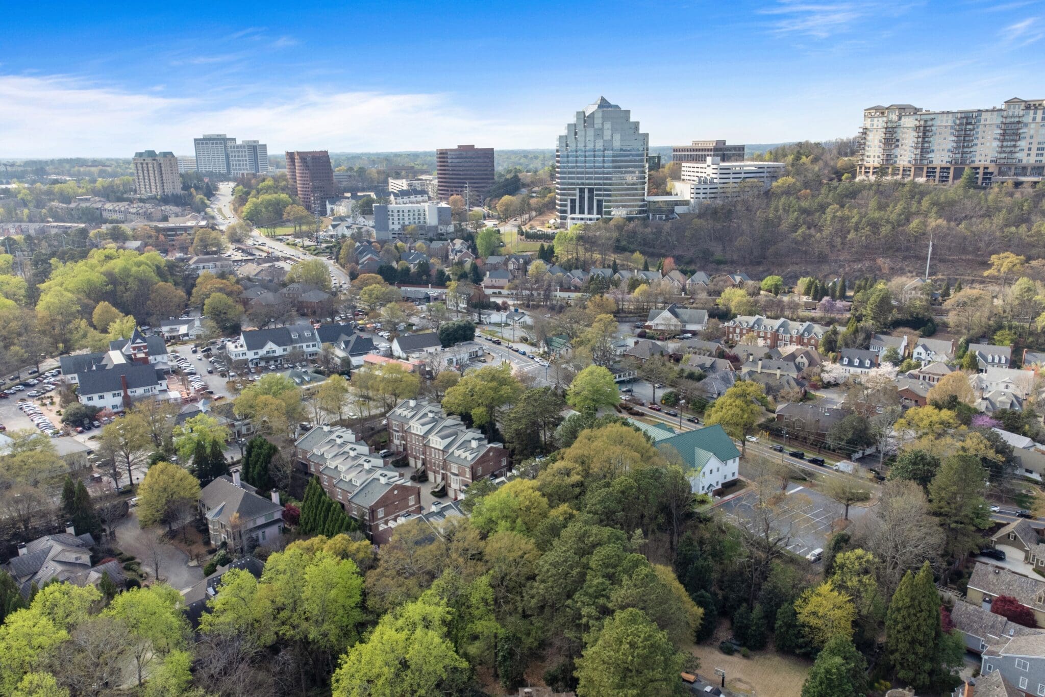 Aerial view of a suburban neighborhood with densely packed houses, surrounding green trees, and various commercial buildings in the background under a clear blue sky.