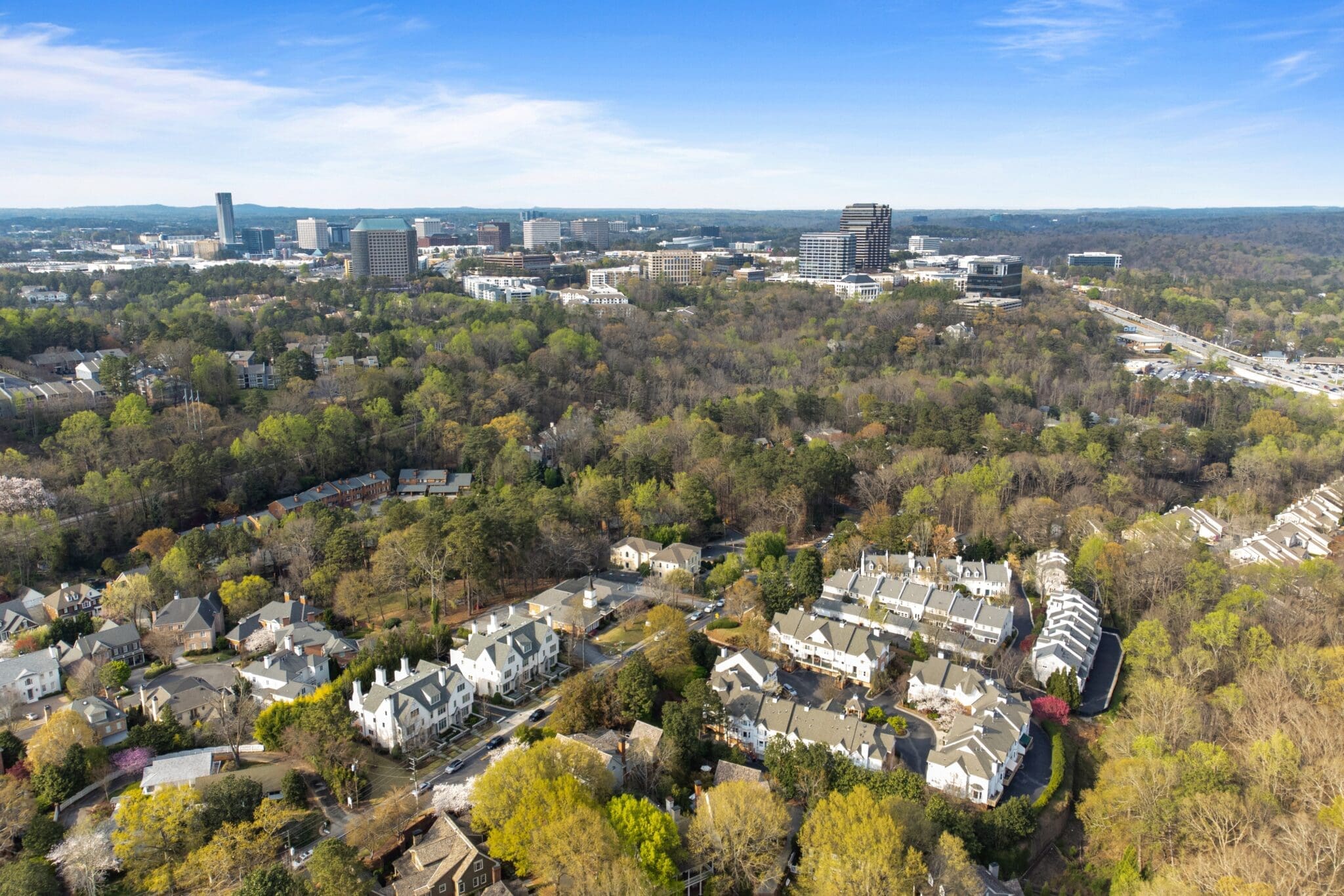 Aerial view of a suburban area with clusters of houses interspersed with trees, leading to a cityscape with multiple tall buildings in the background under a blue sky.