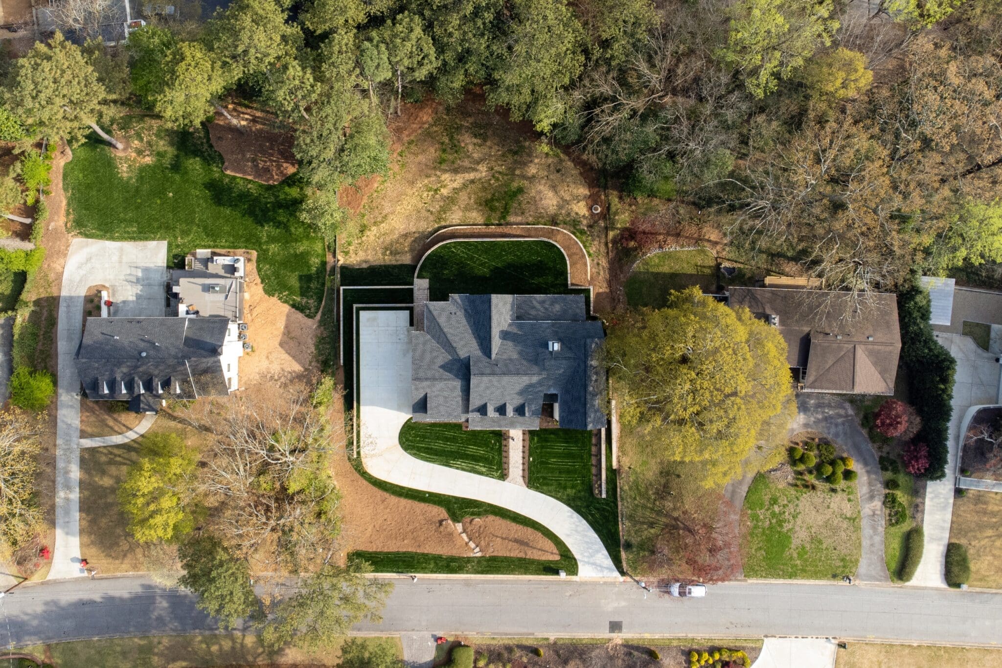 Aerial view of a residential area featuring three houses surrounded by trees with driveways connecting to a main road. One house has a distinct U-shaped driveway.
