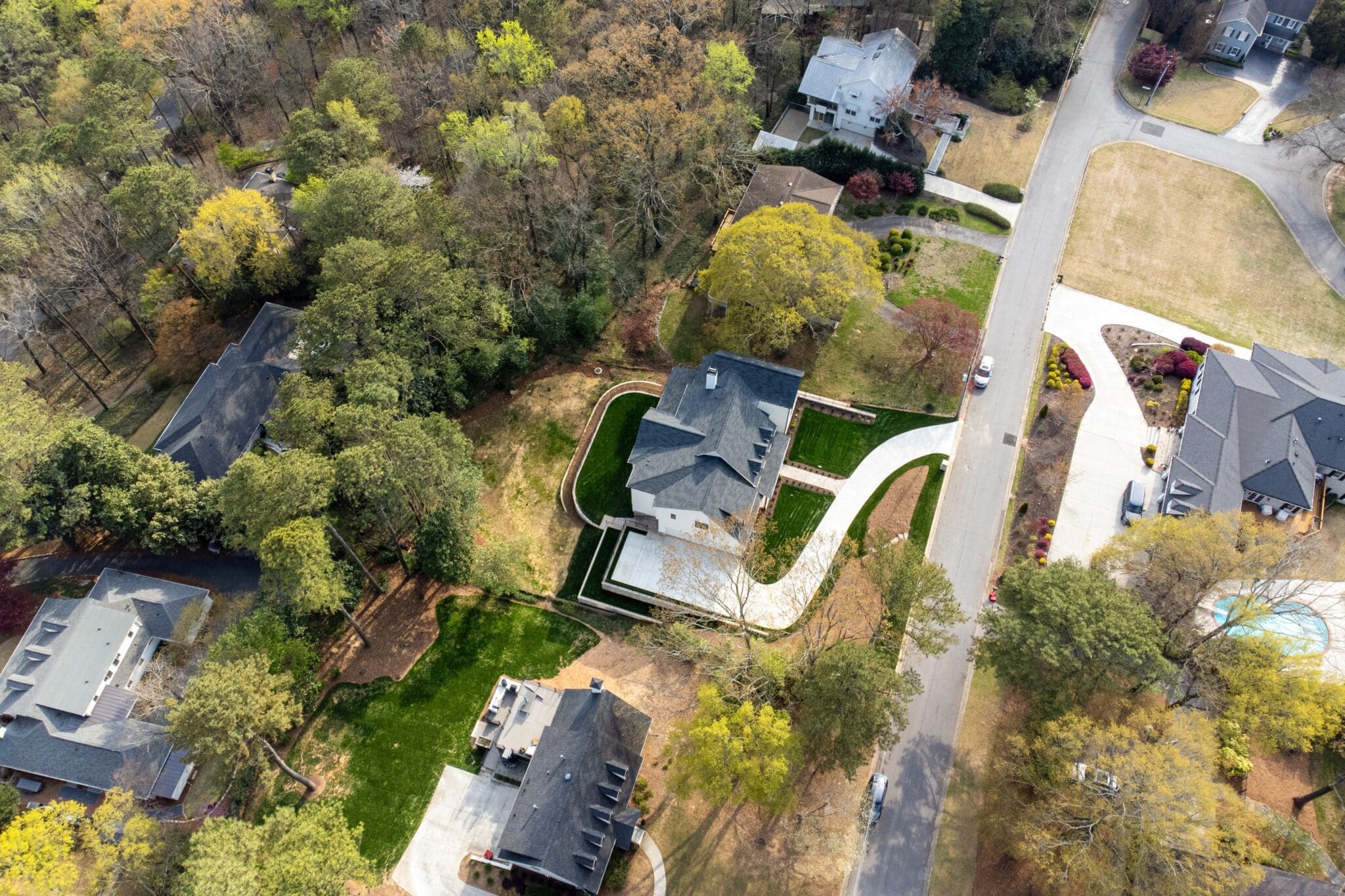 Aerial view of a suburban neighborhood with houses surrounded by trees. A curving driveway leads to a central house, with other homes and roads visible amidst greenery.