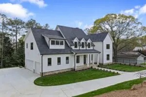 Modern two-story house with white siding, dark roof, multiple gables, large windows, and a two-car garage. It is situated on a landscaped plot with a newly paved driveway and surrounded by trees.