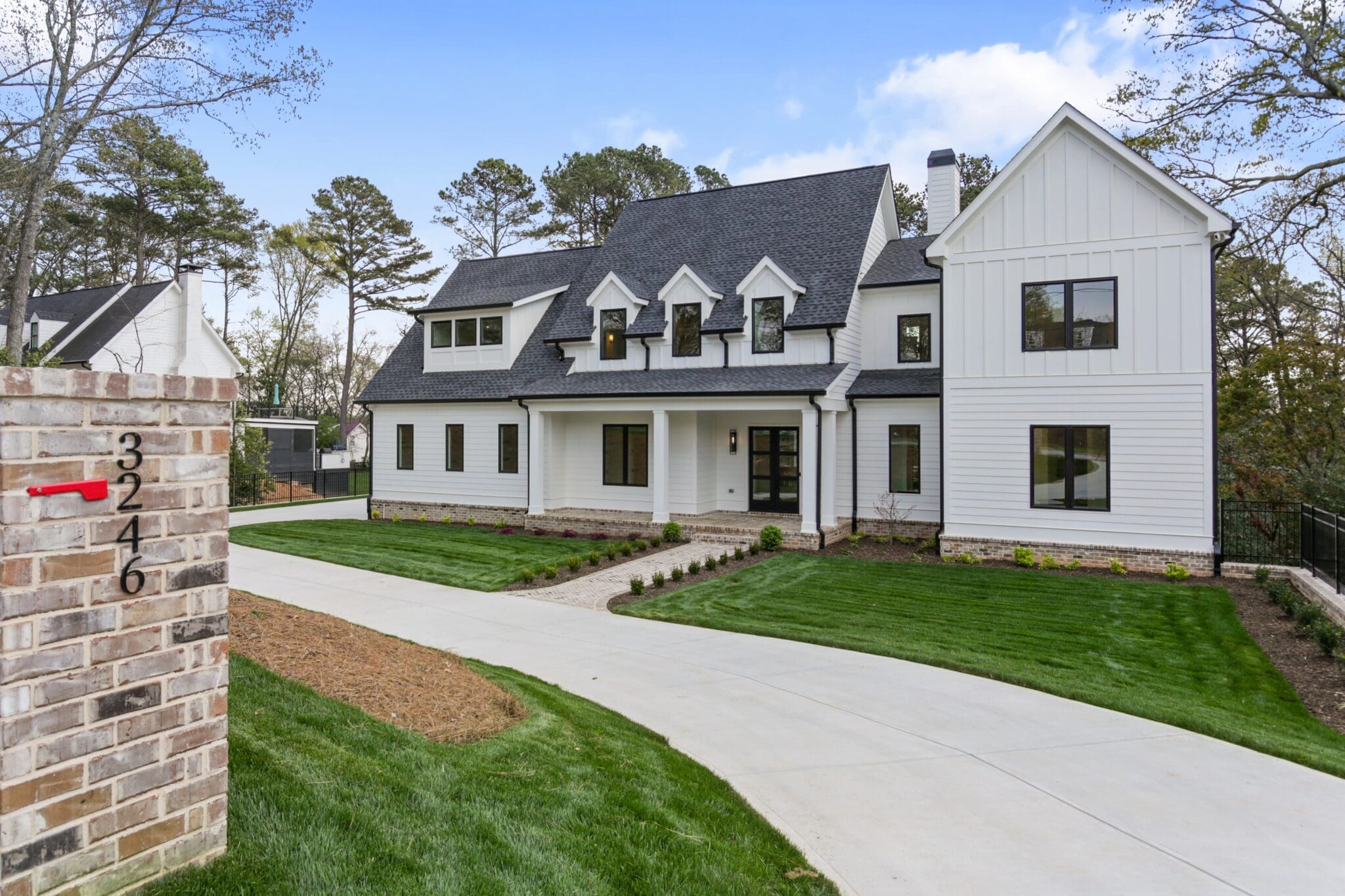 A modern two-story white house with a gabled roof and dormer windows stands behind a brick pillar numbered 3246. The property features a curved driveway and a well-manicured lawn.