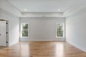 An empty room with light gray walls, a tray ceiling, hardwood floors, and two windows with wooded views.