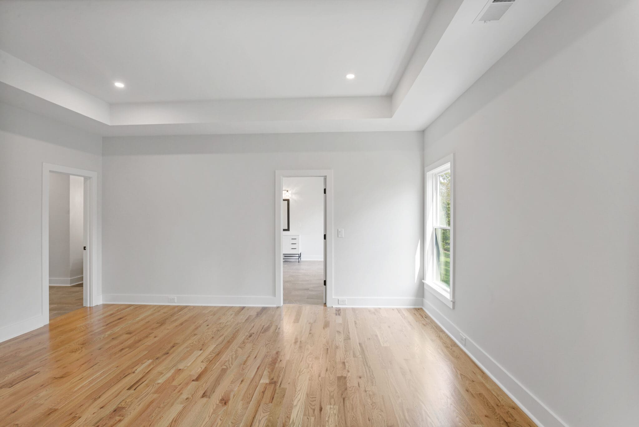 A bright, empty room with light wood flooring, neutral-colored walls, a single window, and two doorways leading to adjacent rooms.