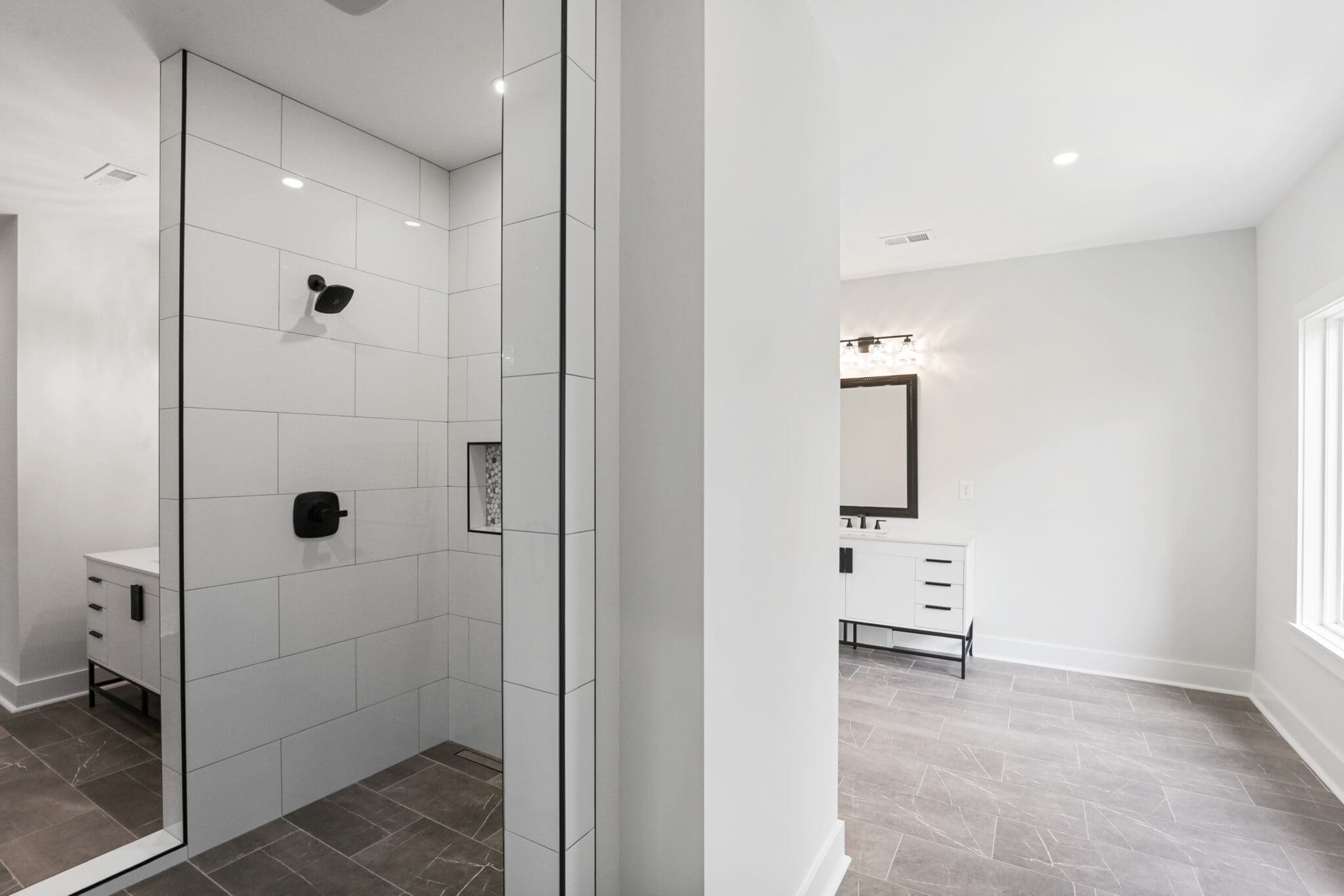 A modern bathroom with a glass-enclosed shower featuring white tiles and black fixtures, adjacent to a vanity with a large mirror, set on gray floor tiles.