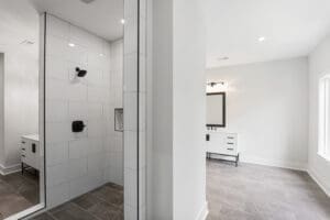 A modern bathroom with a glass-enclosed shower featuring white tiles and black fixtures, adjacent to a vanity with a large mirror, set on gray floor tiles.