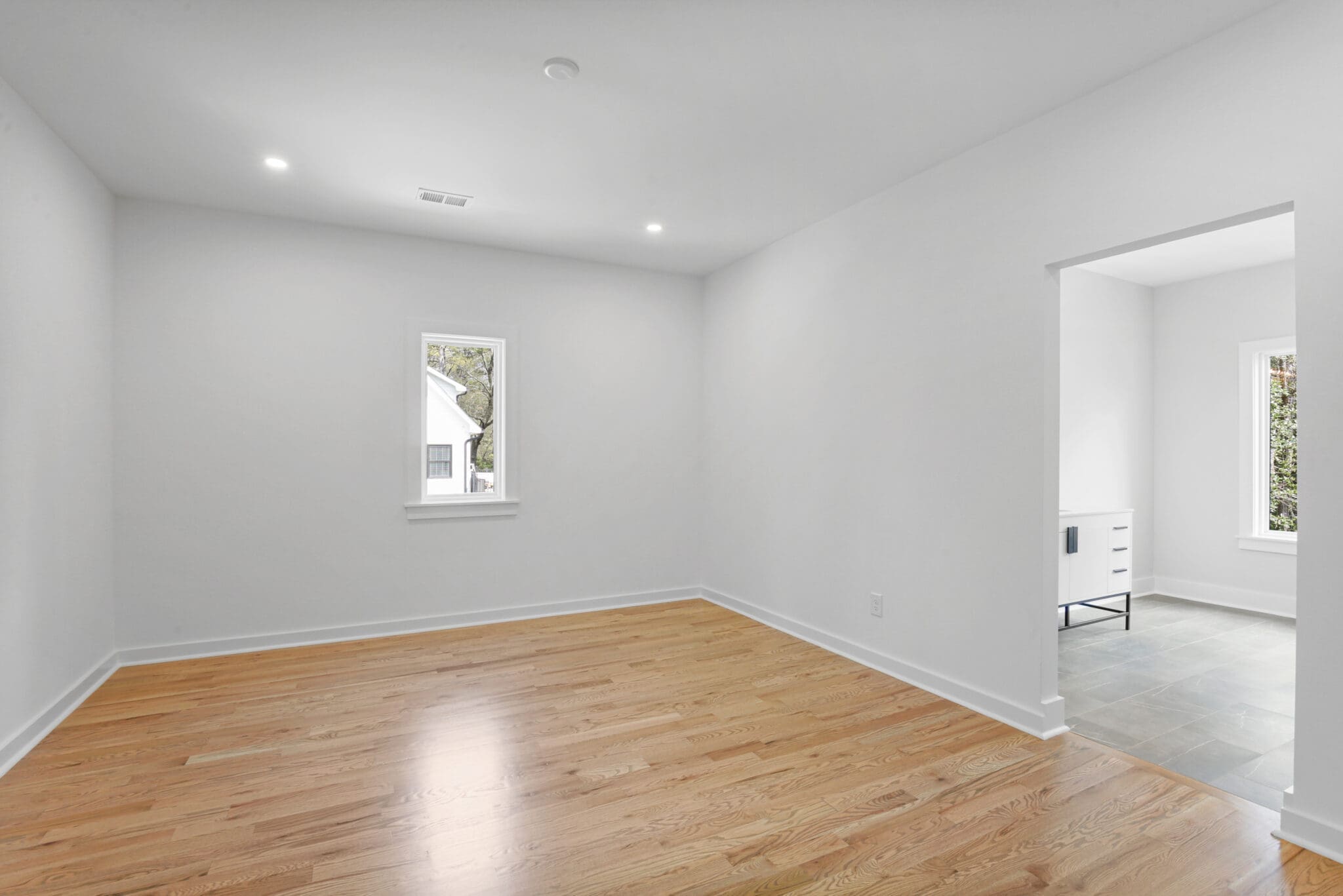 A spacious, unfurnished room with light-colored hardwood floors, a small window, and an open doorway leading to a tiled area with a vanity against the wall.