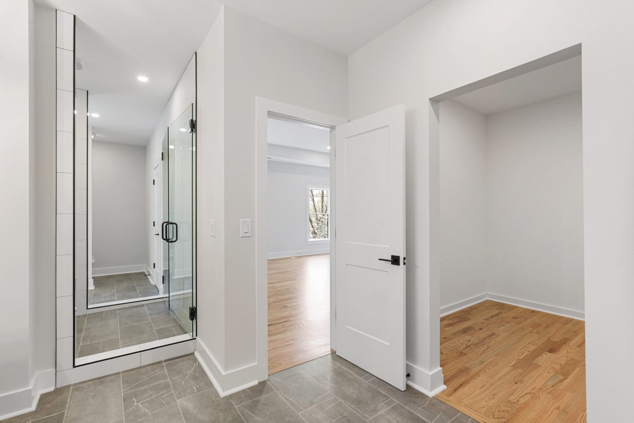 A view of a modern, minimalistic hallway with a glass-enclosed shower, an open door leading to a brightly lit room with hardwood flooring, and an empty alcove.
