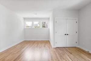 An empty room with light wood flooring, white walls, a small three-pane window, and a double door closet with white doors and black handles.