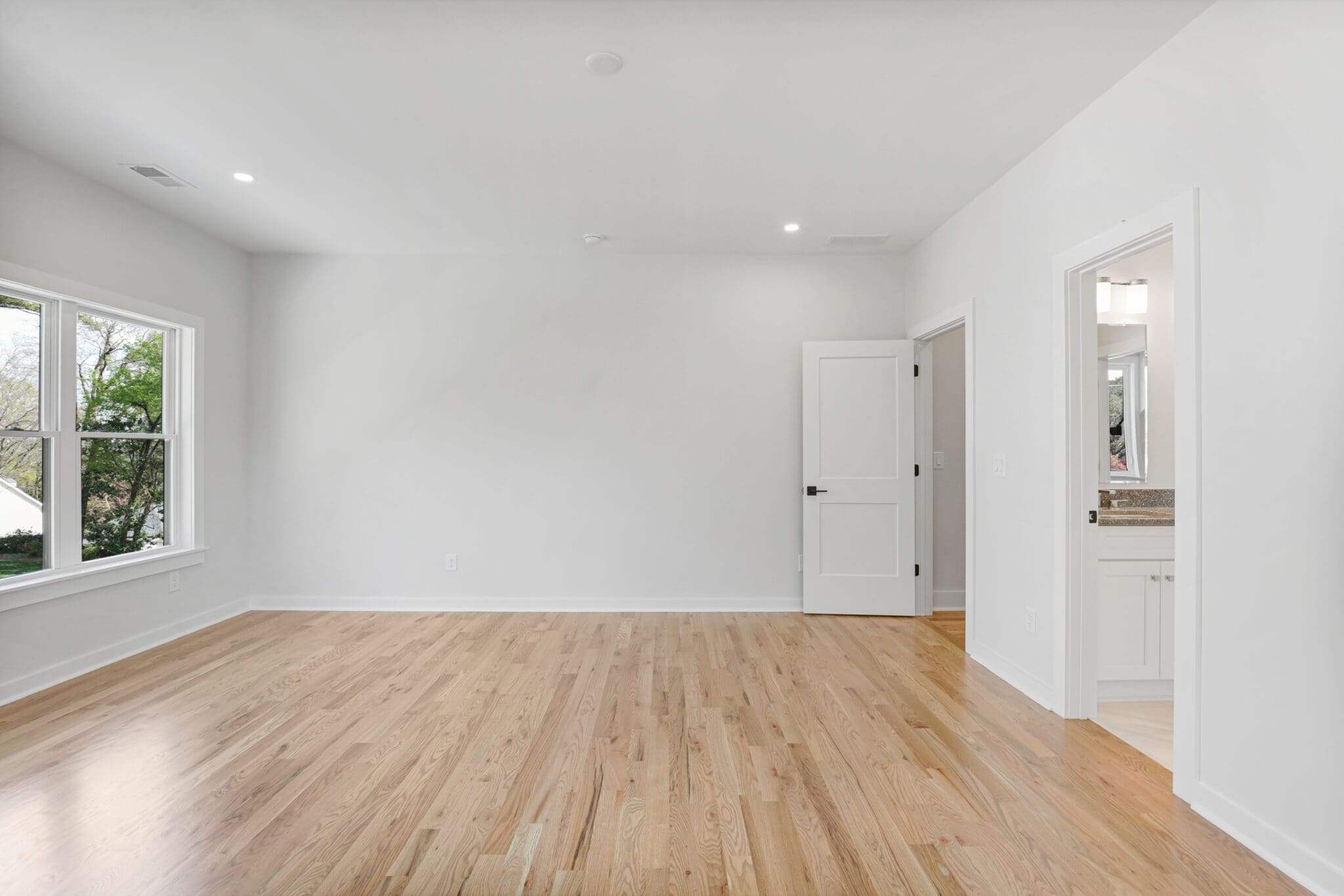 An empty room with light wood floors, white walls, a large window, and a doorway leading to a bathroom with a sink and mirror.