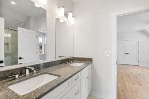 A modern bathroom features a double sink vanity with a speckled countertop, white cabinets, and a large wall mirror. The room is well-lit with light fixtures above the mirror and an adjacent open doorway.