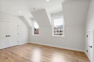 A bright, empty room with hardwood floors, two large windows, angled ceilings, and a pair of closed double doors.
