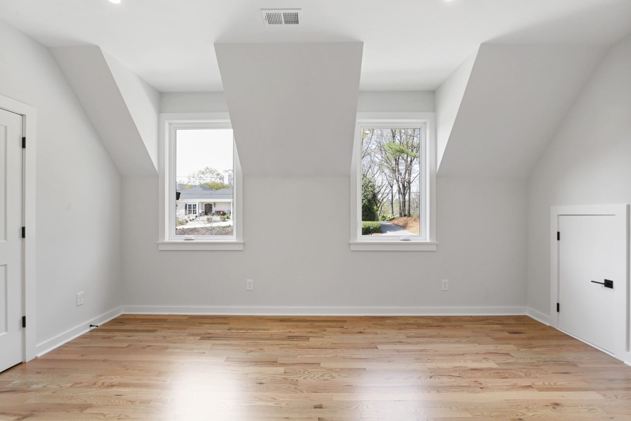 An unfurnished room with white walls, two windows, and wooden flooring. The ceiling features two sloped sections, and there is a small door on the right side of the room.