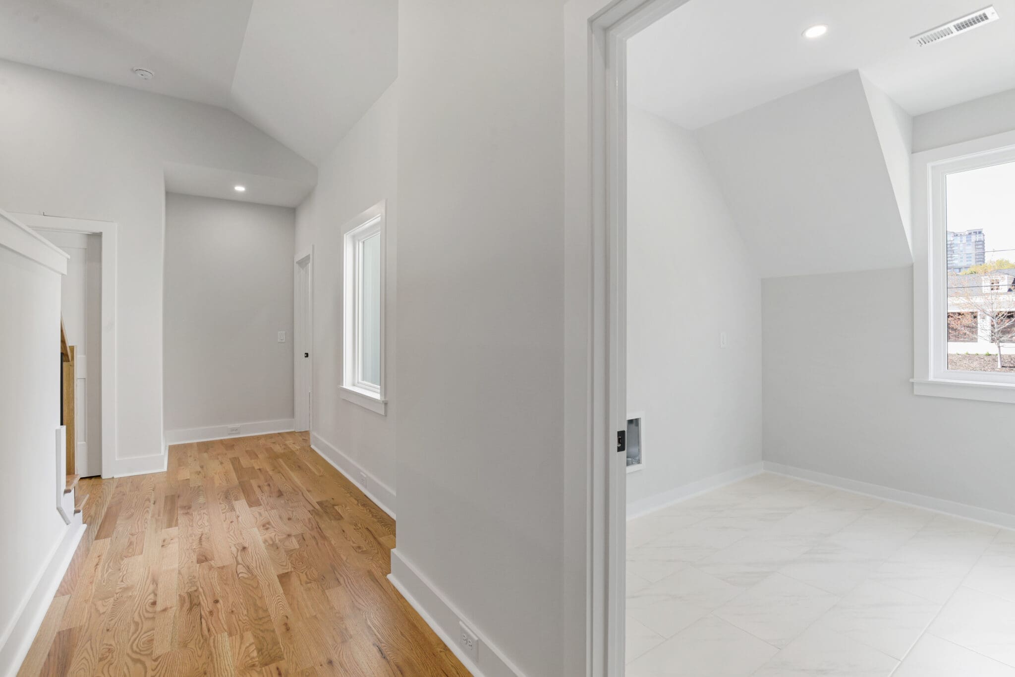 A modern, light-filled hallway with wood flooring leads to a bright room with white tiled flooring. The space features white walls and large windows.