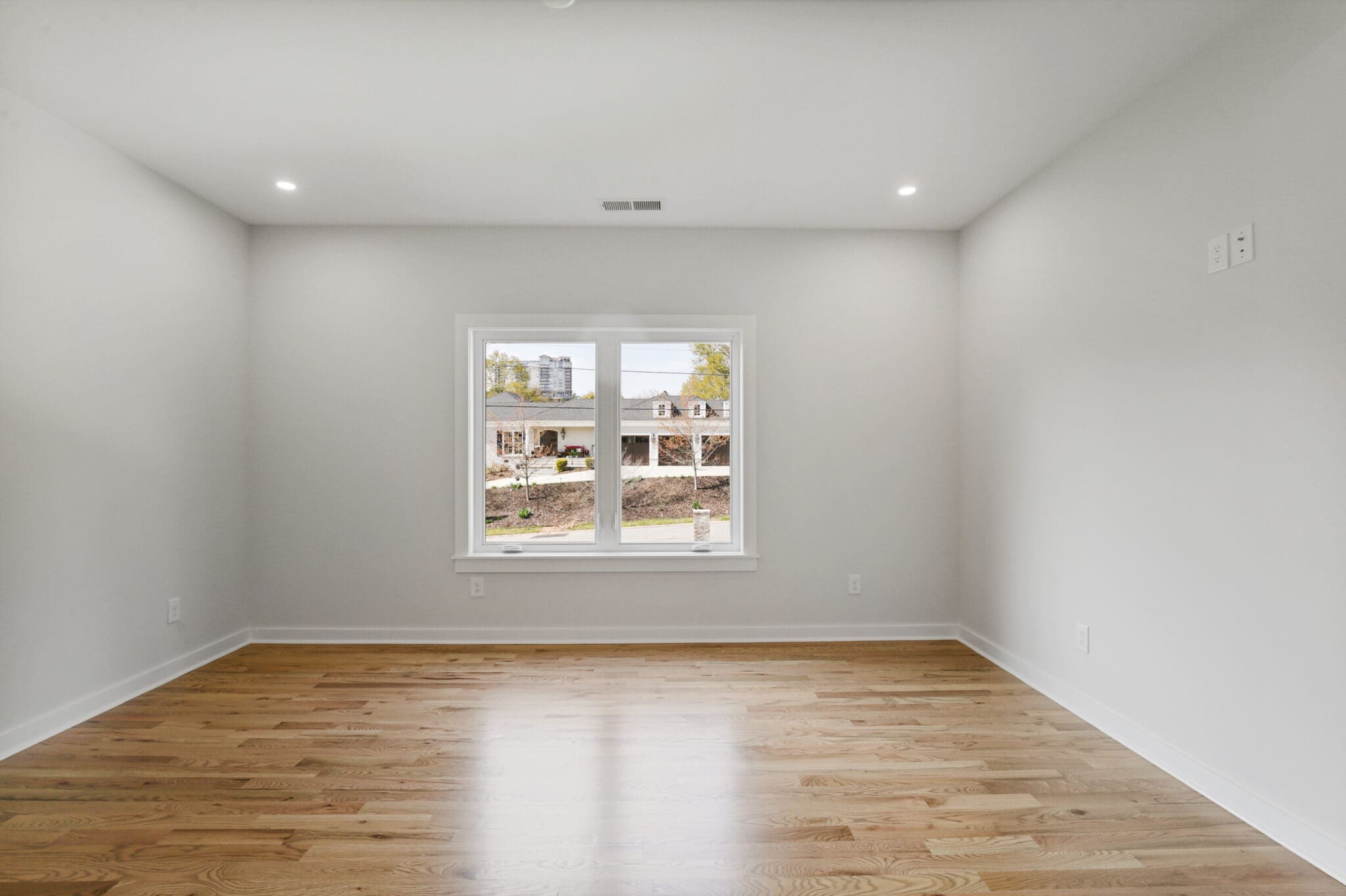 A bright, empty room with light-colored hardwood flooring, white walls, and a large window offering a view of buildings and greenery outside. Ceiling lights are turned on.