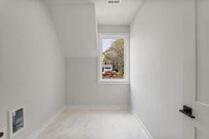 Small, empty room with light gray walls and white tiled floor. A single window shows a view of houses and greenery outside. The room has a vent and a door partially visible on the right.