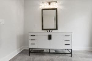 A modern bathroom vanity with a large rectangular mirror above a double drawer cabinet, illuminated by three-bulb light fixtures. The floor is covered with gray tiles.