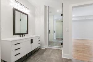 Modern bathroom with a large mirror above a white double-sink vanity, a glass-enclosed shower, grey floor tiles, and beige walls. The room extends into a space with wooden flooring.