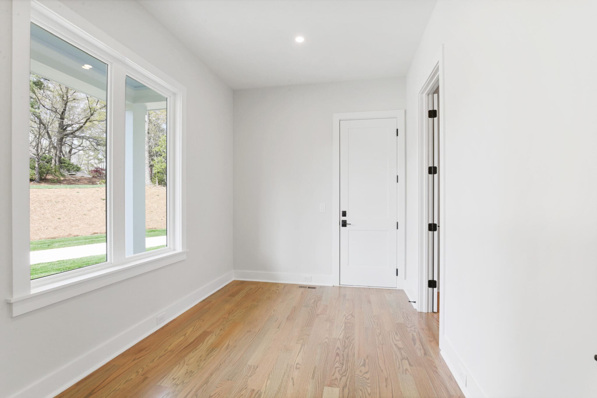 A bright room with light-colored walls, wooden flooring, a large window on the left, and a closed white door on the right.