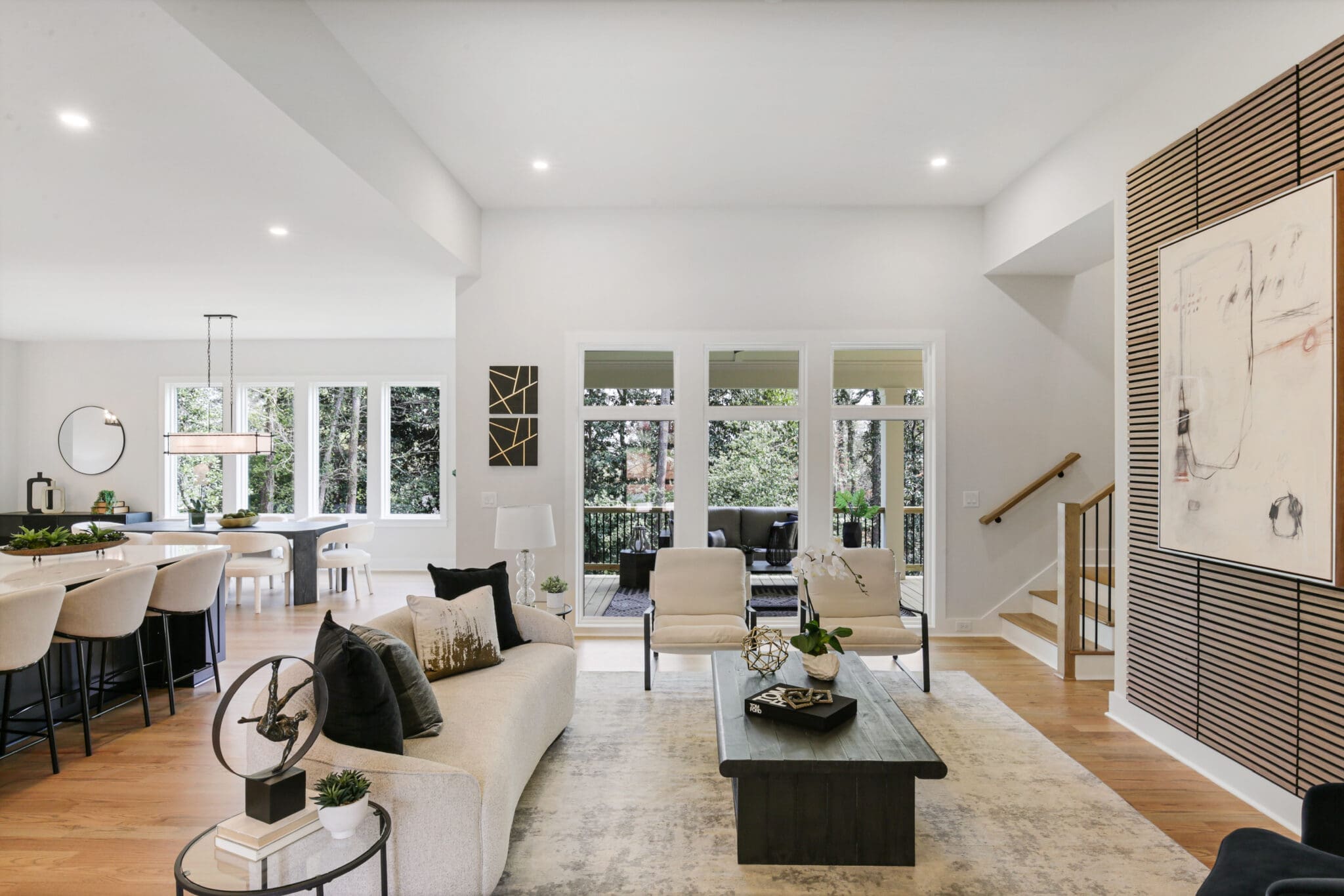 A spacious, modern living room with light-colored decor. Large windows overlook greenery, and a dining area is visible in the background. A staircase is to the right near abstract wall art.