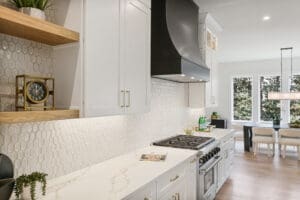 Modern kitchen with white cabinetry, black range hood, hexagonal tile backsplash, and a gas stove. Open shelving holds decor items, and a dining area with a table and chairs is visible in the background.