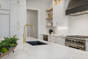 Modern kitchen with white cabinetry, a black sink, gold faucet, and a stainless steel stove. Marble countertop with decorative items and open shelves. Bright lighting and a tiled backsplash.