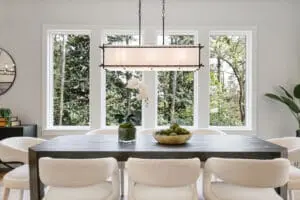 A modern dining room features a rectangular wooden table with six white upholstered chairs, a potted plant centerpiece, a bowl of green decorative balls, and a large window with a view of trees.