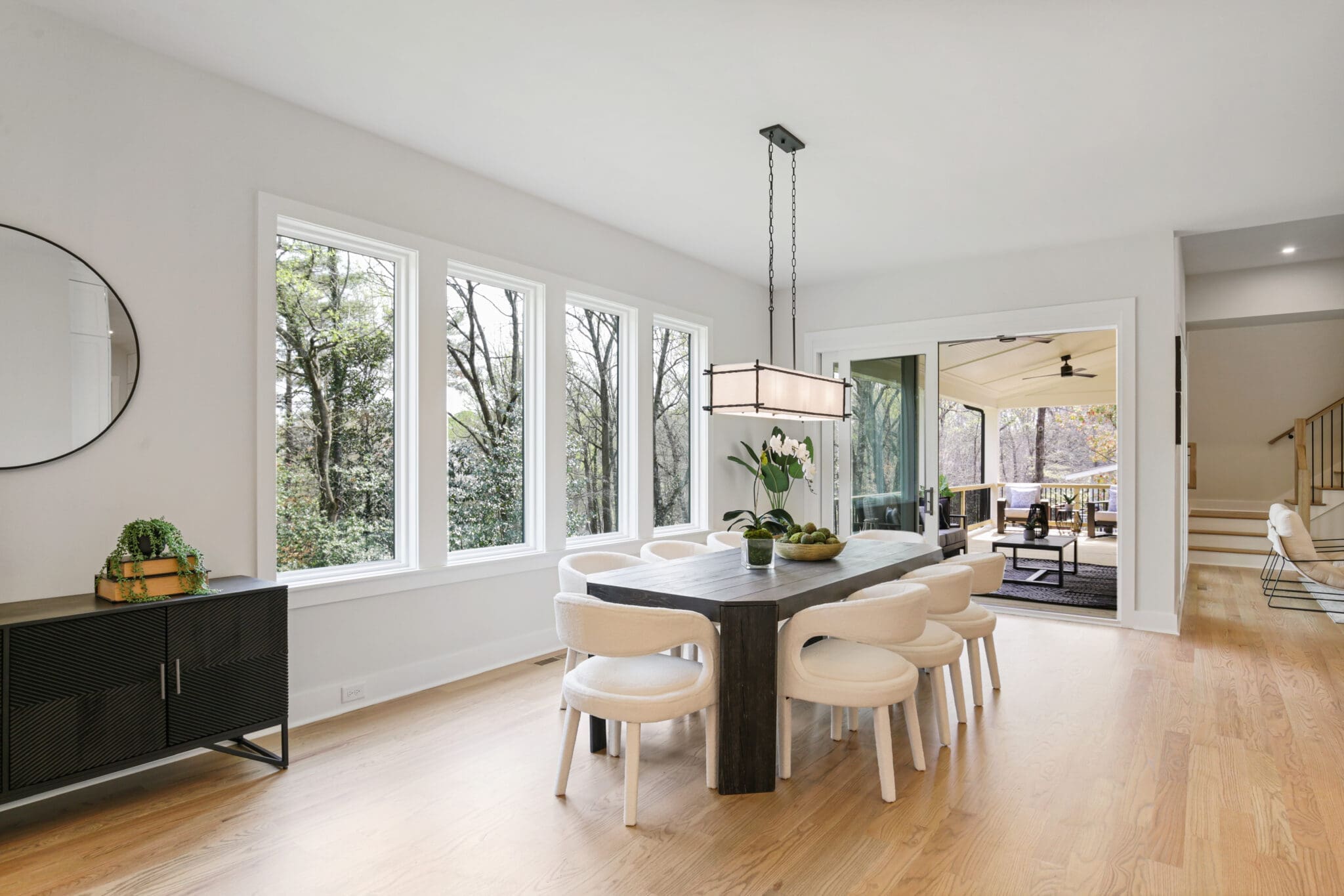 A modern dining area with a large wooden table and eight white chairs, next to wide windows and a sliding door opening to a spacious outdoor patio.
