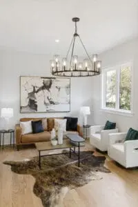 A modern living room with a brown sofa, two white chairs, abstract art, a geometric chandelier, and a cowhide rug. Two lamps flank the sofa, and a coffee table with decor sits in front.