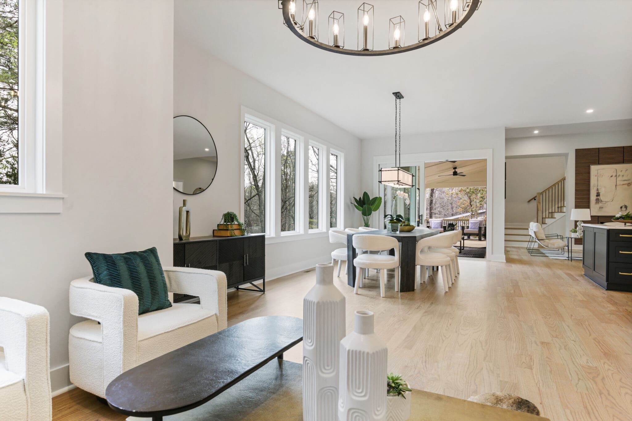A bright, modern open-concept living and dining area with light wood flooring, white furniture, large windows, and a contemporary chandelier. A kitchen is visible in the background.