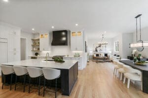 Spacious modern kitchen with a large island, white cabinetry, and black accents. The dining area and living room are visible in the background. Hardwood floors and contemporary light fixtures throughout.