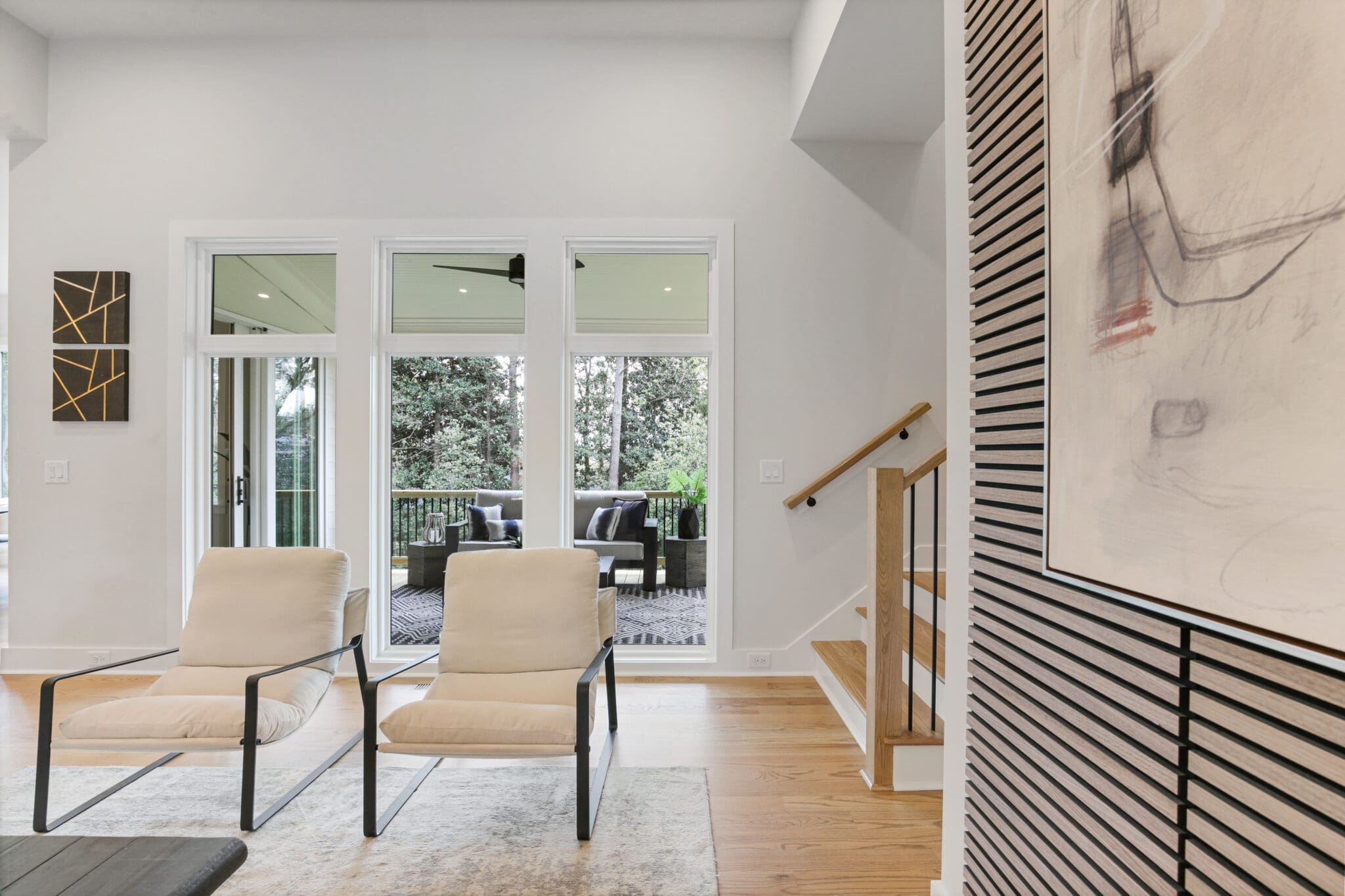 A modern, open living space with two beige chairs facing glass doors leading to an outdoor patio. Art pieces adorn the walls, and a wooden staircase is visible to the right.