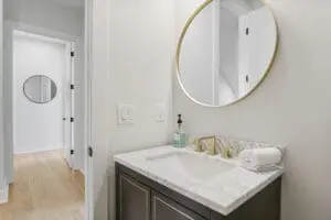 Modern bathroom with a marble countertop sink, brass faucet, round mirror, and towel. A hallway with another mirror is visible through the open door.