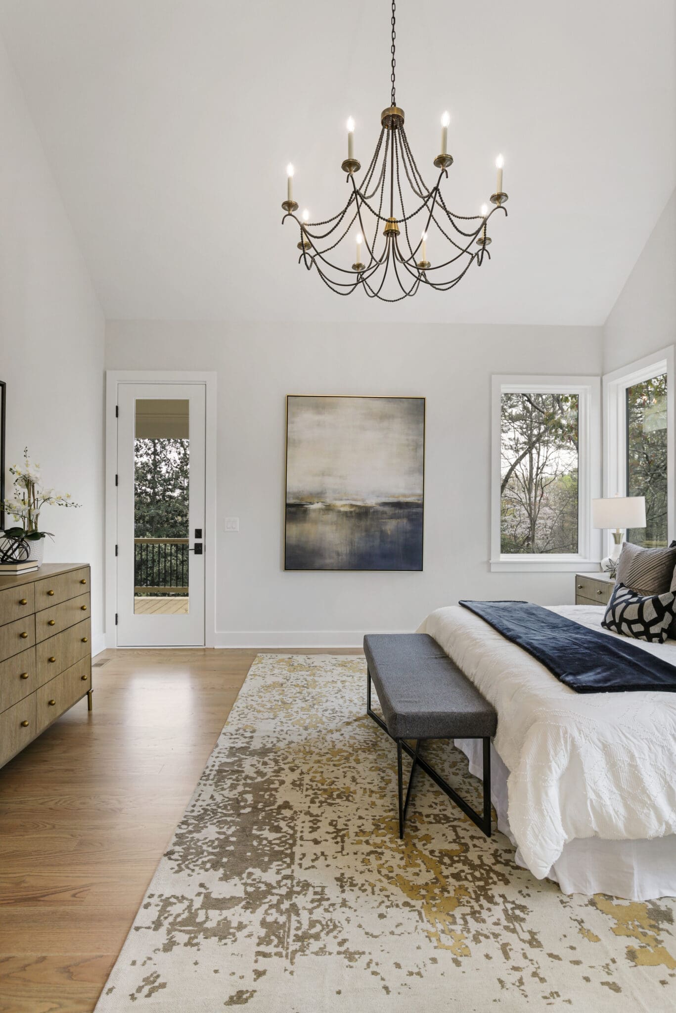 A modern bedroom features a large bed with white bedding, a bench at the foot, a dresser, and a chandelier. Two windows and a door provide natural light. A painting hangs on a wall above the bed.