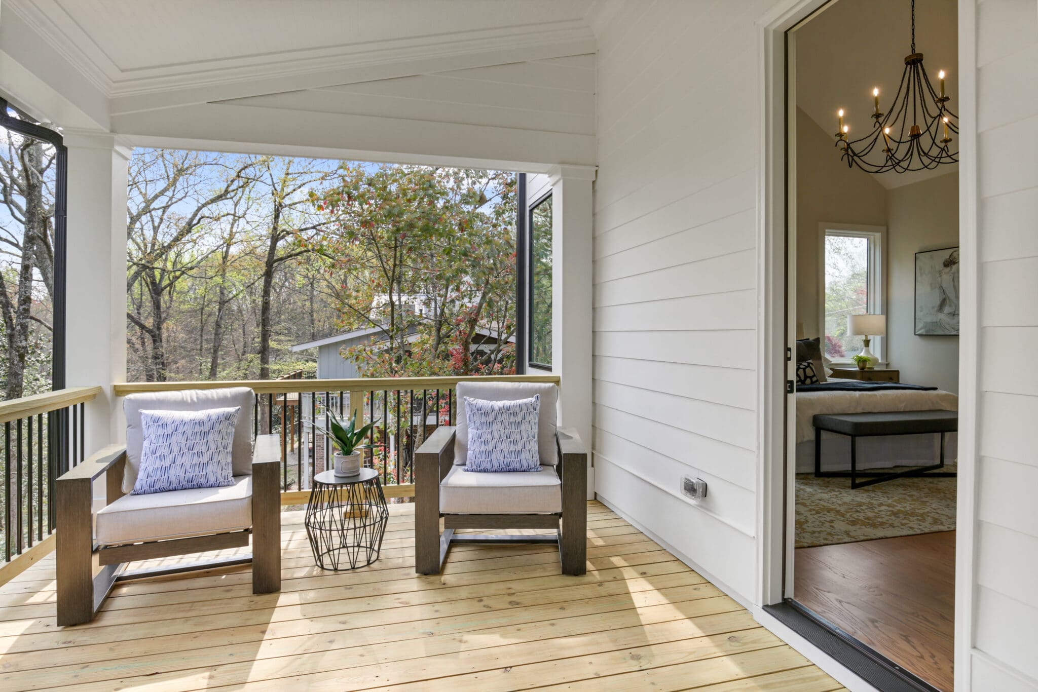 A wooden deck with two cushioned chairs and a small table with a plant. The deck overlooks trees and a neighboring house. Open door shows a bedroom with a chandelier and a bed.