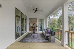 Covered porch with a ceiling fan, modern furniture, a patterned rug, and glass sliding doors leading to an interior space. The porch overlooks a green, tree-filled outdoor area.