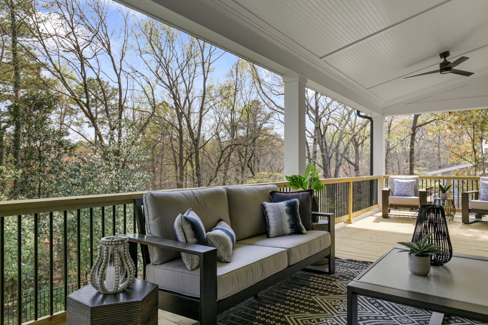 A covered outdoor patio with forest views features a cushioned sofa, patterned rug, potted plants, and a ceiling fan. Additional seating and a table are visible in the background.