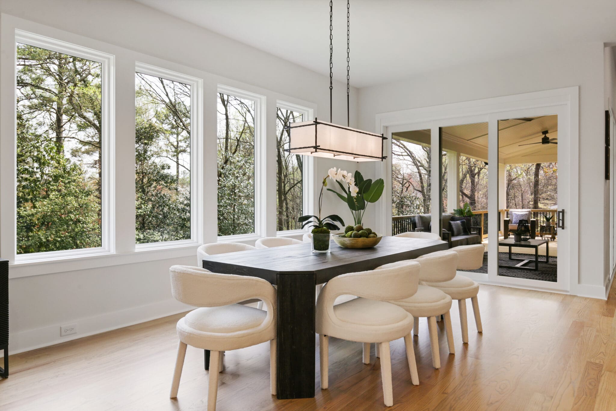 Modern dining area with a rectangular table, eight white chairs, a potted orchid centerpiece, and large windows providing a view of a sunlit patio and trees outside.
