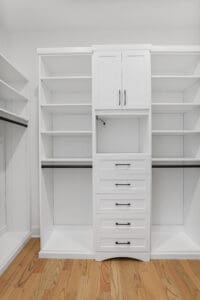 A white closet system with wooden flooring includes open shelves, a cabinet with doors, a section with a hanging rod, and multiple closed drawers with black handles.