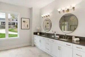 Modern bathroom with double vanity, white cabinets, black countertop, two round mirrors, and wall-mounted light fixtures. A window on the left allows natural light. Wall art and decor accents are present.