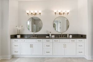 A double vanity bathroom with two round mirrors, dual sinks, white cabinets, and a dark countertop, illuminated by two modern light fixtures above. A potted orchid and toiletries are on the counter.