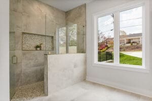 A modern bathroom features a walk-in shower with a glass enclosure, mosaic tile accents, and a large window overlooking a suburban neighborhood.