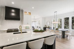 Modern kitchen and dining area with marble countertops, white cabinets, black range hood, and a large island. The dining area features a rectangular table and chairs near large windows.